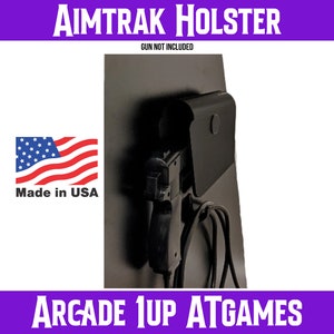 May include: Black plastic gun holster mounted on a black surface. The holster is labelled "Aimtrak Holster" and "Gun Not Included". The text "Arcade 1Up AtGames" is at the bottom of the image. A small American flag with the text "Made in USA" is in the lower left corner of the image.