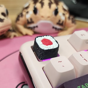 May include: A sushi-shaped keycap sits on a light pink keyboard. The keycap is black with white rice and a red center, resembling a maki roll. Two gecko figurines are in the background.