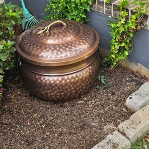 May include: A round, copper-colored metal garden hose storage pot with a hammered texture. The lid features a decorative gold-toned handle shaped like a small branch with two leaves. The container has a wide, circular base and a matching lid.