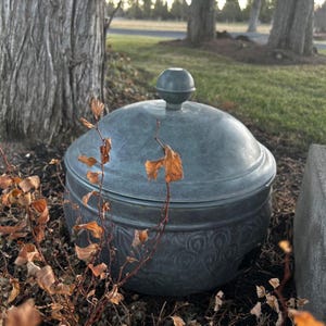 May include: A round, dark grey metal container with a domed lid and a spherical knob handle. The exterior surface features a subtle, embossed repeating pattern of curved lines and shapes, finished with a weathered, matte patina texture.
