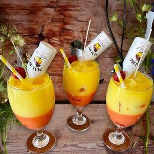 May include: Three yellow and orange cocktails with a layer of yellow, then orange, then a layer of yellow again. Each cocktail is in a clear glass with a stem and a round base. Each cocktail has a straw in it and a small bottle of Malibu Coconut Rum on top. The bottles are white with black text that reads 