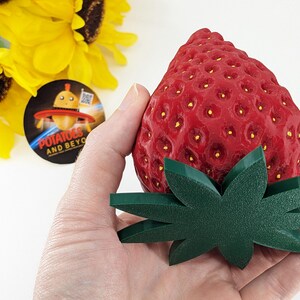May include: A large, red strawberry with a textured surface and small yellow seeds, paired with a green leaf base. The strawberry is held in a hand, with sunflowers and a circular logo in the background.