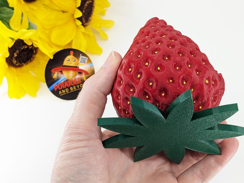 May include: A large, red strawberry with a textured surface and small yellow seeds, paired with a green leaf base. The strawberry is held in a hand, with sunflowers and a circular logo in the background.