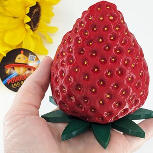May include: A large, red strawberry sculpture with a textured surface and small yellow seeds. The strawberry rests on a green, leaf-shaped base. The sculpture is held in a hand, with a sunflower and a sticker in the background.