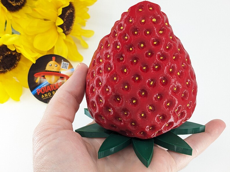 May include: A large, red strawberry sculpture with a textured surface and small yellow seeds. The strawberry rests on a green, leaf-shaped base. The sculpture is held in a hand, with a sunflower and a sticker in the background.