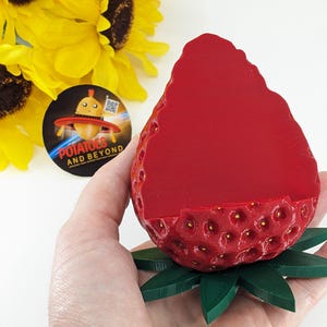 May include: A red strawberry-shaped container with a green leaf base. The container has a textured surface with small yellow dots. A round sticker with the text "POTATOES AND BEYOND" is visible in the background.