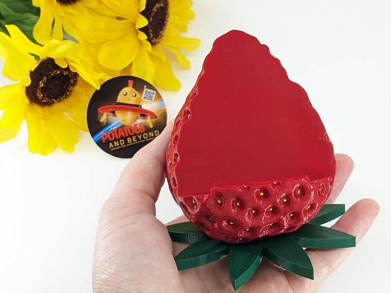 May include: A red strawberry-shaped container with a green leaf base. The container has a textured surface with small yellow dots. A round sticker with the text "POTATOES AND BEYOND" is visible in the background.