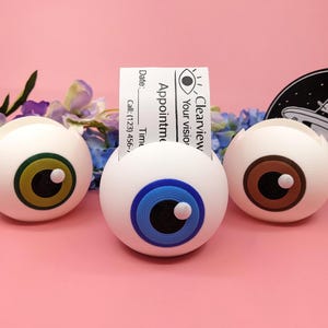 May include: Three white eyeball-shaped desk organizers with different colored irises: green, blue, and brown. A business card with the text "Clearview Your vision" is in the center organizer. A black and white graphic is in the background.