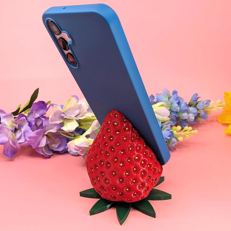 May include: A vibrant red strawberry-shaped phone stand with a blue phone resting on it. The stand has green leaves at the base. The background features purple and blue flowers against a pink backdrop.