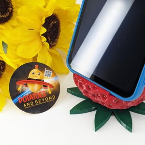 May include: A smartphone in a blue case rests on a red strawberry-shaped phone stand with green leaves. A round sticker with the text "Potatoes and Beyond" and a cartoon potato in a UFO is next to yellow sunflowers.