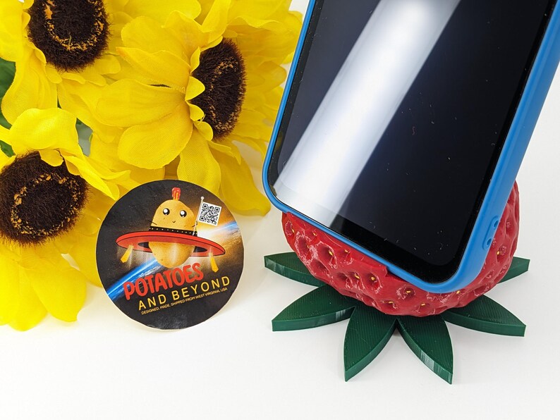 May include: A smartphone in a blue case rests on a red strawberry-shaped phone stand with green leaves. A round sticker with the text "Potatoes and Beyond" and a cartoon potato in a UFO is next to yellow sunflowers.
