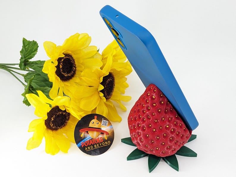 May include: A blue phone case rests on a red strawberry-shaped phone stand with green leaves. Yellow sunflowers and a sticker with the text "Potatoes and Beyond" are also in the image.