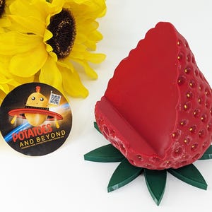 May include: A bright red strawberry-shaped phone holder with a green leaf base. The holder has a textured surface with small yellow seeds. A round sticker with the text "Potatoes and Beyond" and a cartoon potato in a UFO is also visible.