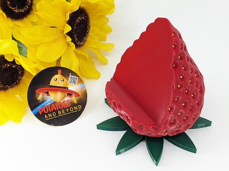 May include: A bright red strawberry-shaped phone holder with a green leaf base. The holder has a textured surface with small yellow seeds. A round sticker with the text "Potatoes and Beyond" and a cartoon potato in a UFO is also visible.