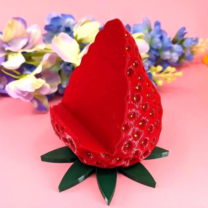 May include: A red strawberry-shaped phone stand with a green leaf base. The stand has a textured surface with yellow seed details. The background features pink and purple flowers.