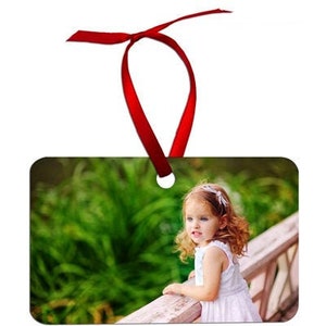 May include: Rectangular ornament with a photo of a young girl in a white dress, with a red ribbon for hanging. The girl is leaning on a wooden railing with a green background.