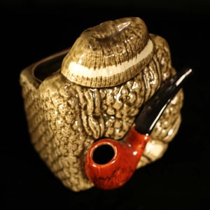 May include: A ceramic planter shaped like a person wearing a brown hat and holding a red pipe. The planter has a textured surface and is a light brown colour.