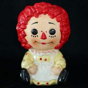 May include: A ceramic doll with red curly hair, large black eyes, and a red nose and lips. The doll wears a yellow dress with red polka dots and a white apron. Black shoes complete the look. The doll is a vintage collectible.