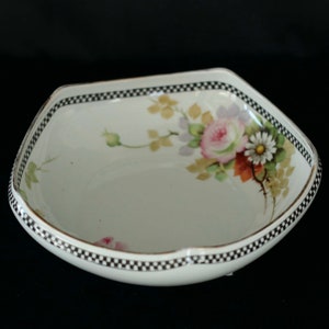 Peut inclure: Un plat en porcelaine blanc de forme carrée avec un motif floral. Le plat présente une bordure à carreaux noirs et blancs et une bordure dorée. Le motif floral comprend des roses roses, des marguerites blanches et des feuilles vertes. Fond noir.