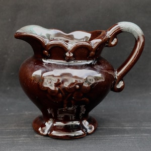 May include: A brown ceramic creamer with a scalloped rim and a handle. The creamer has a glossy finish and is decorated with raised floral designs.