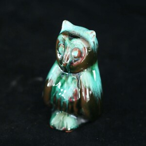 Könnte beinhalten: Keramik-Eulenfigur mit glänzendem Finish. Die Eule ist hauptsächlich grün und braun, mit einem getropften Glasureffekt. Die Figur hat große, runde Augen und einen kleinen Schnabel, vor schwarzem Hintergrund.