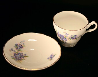 May include: A white teacup and saucer with a floral pattern of blue forget-me-nots and green leaves. The edges of the cup and saucer are trimmed with gold.