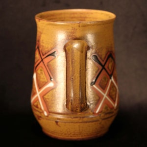 Puede incluir: Una taza de cer&aacute;mica marr&oacute;n con un dise&ntilde;o geom&eacute;trico en tonos naranja, blanco y negro. La taza tiene un asa y una forma redondeada.
