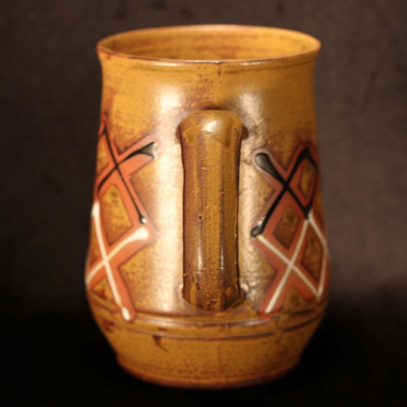 Puede incluir: Una taza de cer&aacute;mica marr&oacute;n con un dise&ntilde;o geom&eacute;trico en tonos naranja, blanco y negro. La taza tiene un asa y una forma redondeada.