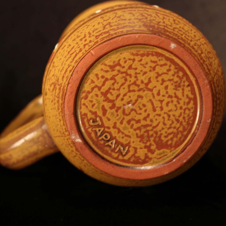 Puede incluir: Una taza de cer&aacute;mica marr&oacute;n con una superficie texturizada y un borde rojo. La parte inferior de la taza tiene la palabra "JAPAN" impresa en dorado.