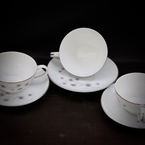 Puede incluir: Cuatro tazas de té y platillos blancos. Cada taza tiene un asa y se asienta sobre un platillo a juego. Los platillos y las tazas tienen un delicado estampado floral en gris y rosa, con un borde plateado. El fondo es negro.