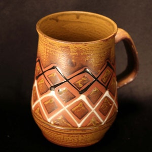 Puede incluir: Una taza de cer&aacute;mica marr&oacute;n con un patr&oacute;n geom&eacute;trico en tonos marrones, rojos, blancos y negros. La taza tiene un asa y una base redondeada.
