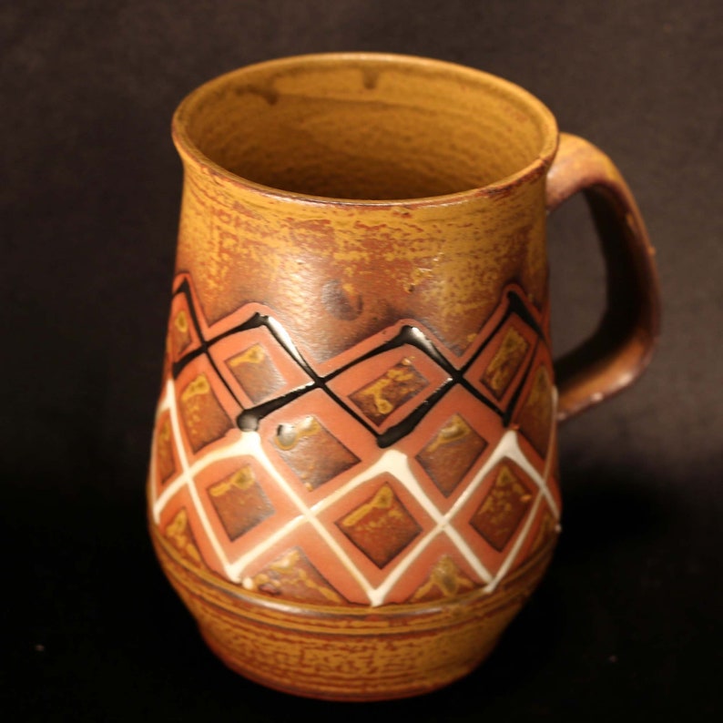 Puede incluir: Una taza de cer&aacute;mica marr&oacute;n con un patr&oacute;n geom&eacute;trico en tonos marrones, rojos, blancos y negros. La taza tiene un asa y una base redondeada.