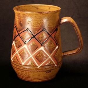 Puede incluir: Una taza de cer&aacute;mica marr&oacute;n con un dise&ntilde;o geom&eacute;trico en negro, blanco y rojo. La taza tiene una base ancha y un asa redondeada.
