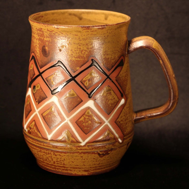 Puede incluir: Una taza de cer&aacute;mica marr&oacute;n con un dise&ntilde;o geom&eacute;trico en negro, blanco y rojo. La taza tiene una base ancha y un asa redondeada.