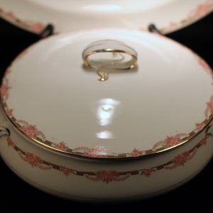 May include: White porcelain serving dish with lid and handles. The dish is decorated with a pink floral pattern and gold trim. The lid has a gold handle. A matching plate is visible in the background.