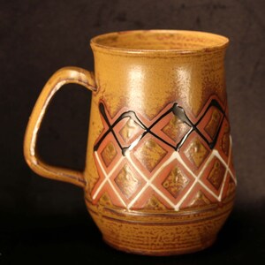 Puede incluir: Una taza de cer&aacute;mica marr&oacute;n con un dise&ntilde;o geom&eacute;trico en negro, blanco y rojo. La taza tiene un asa y una forma redondeada.