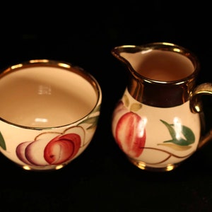 May include: A vintage ceramic sugar and creamer set with a gold trim. The sugar bowl and creamer are decorated with a floral design featuring pink, purple, and green fruit.