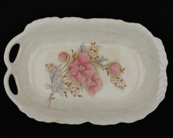 May include: Cream-colored, oval serving dish with a scalloped edge and two handles. The dish features a floral design with pink flowers, blue-gray leaves, and small white blossoms. The dish is ideal for serving appetizers or desserts.