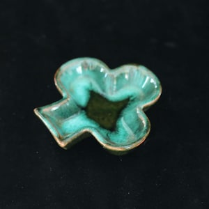 May include: A small, ceramic dish in the shape of a four-leaf clover. The dish has a glossy, teal-green glaze with darker green accents in the center. The edges are a darker brown. The dish is likely used for serving small portions.