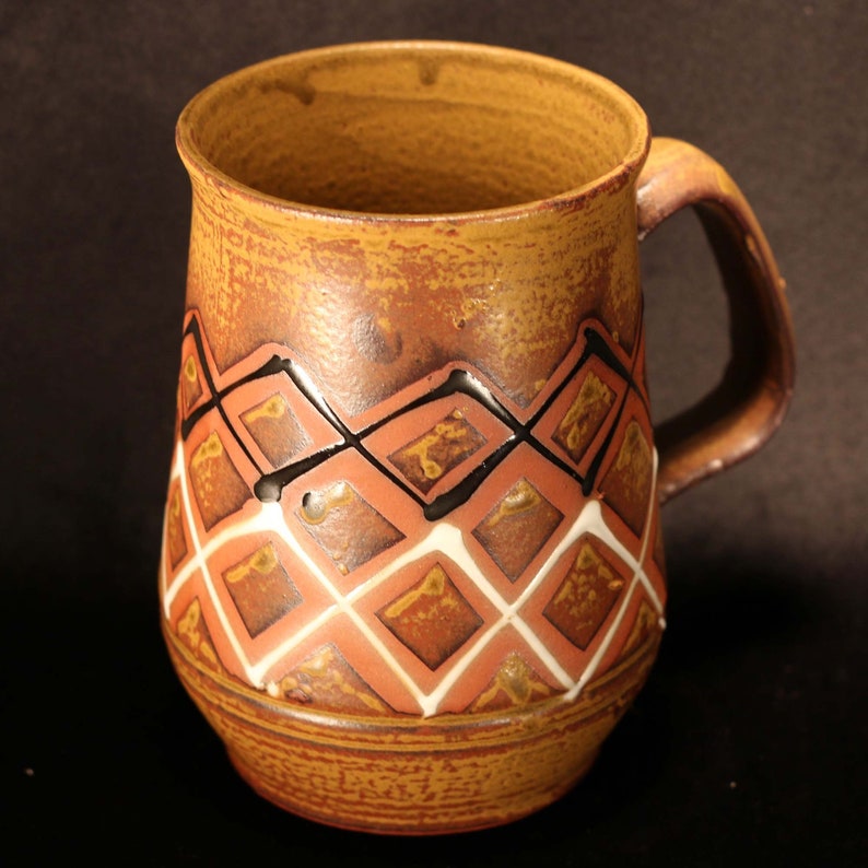 Puede incluir: Una taza de cer&aacute;mica marr&oacute;n con un patr&oacute;n geom&eacute;trico de diamantes rojos, blancos y negros. La taza tiene un asa y un fondo redondeado.
