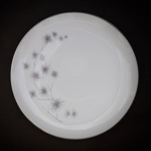 Peut inclure: Une assiette à dîner en céramique blanche avec un bord argenté et un motif floral délicat. Le motif présente des formes en forme d'éclat d'étoile grises avec des centres rouges, disposées le long de fines tiges grises. L'assiette est ronde et repose sur un fond noir.