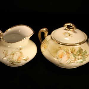 May include: A vintage porcelain creamer and sugar bowl set with a white background and delicate floral designs in shades of yellow, green, and pink. The set features gold accents around the edges.