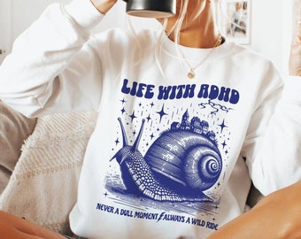 May include: White sweatshirt featuring a blue graphic of a snail carrying a small town on its shell. The text "Life with ADHD" is above the snail and "Never a dull moment, always a wild ride" is below the snail.