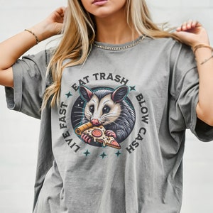 Peut inclure: T-shirt gris surdimensionné avec un motif d'opossum mangeant une pizza. Le motif est entouré d'un cercle avec le texte "Live Fast Eat Trash Blow Cash".