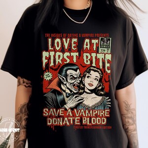May include: A black t-shirt with a vintage comic book style graphic featuring a vampire and a woman with the text "Love at First Bite" and "Save a Vampire Donate Blood Limited Transylvanian Edition".