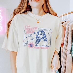 May include: A light yellow t-shirt with a graphic print of a woman with a retro comic book style. The text on the shirt reads "If the planets can align maybe so can I".