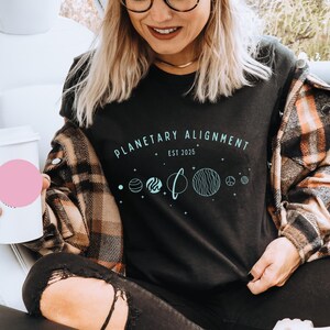 May include: A black t-shirt with the text "PLANETARY ALIGNMENT EST 2025" and a graphic of planets in a line.