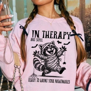 May include: A pale pink sweatshirt featuring the text "IN THERAPY BUT STILL READY TO HAUNT YOUR NIGHTMARES" and a graphic of a raccoon in mummy bandages, bats, and spiderwebs. This is a design from a clothing collection.