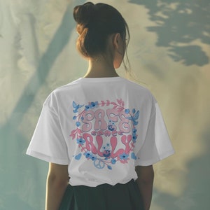 Peut inclure: T-shirt blanc avec un motif graphique rose et bleu qui dit "Safe Space Ally" entouré de fleurs et de feuilles.