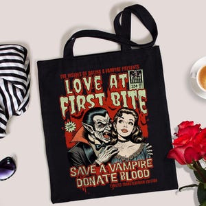 May include: Black tote bag with a comic book style design featuring a vampire and a woman. The text reads "Love at First Bite" and "Save a Vampire Donate Blood".